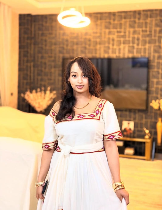 Woman in a white Ethiopian Libs with colorful patterns standing in a room with decorative elements.