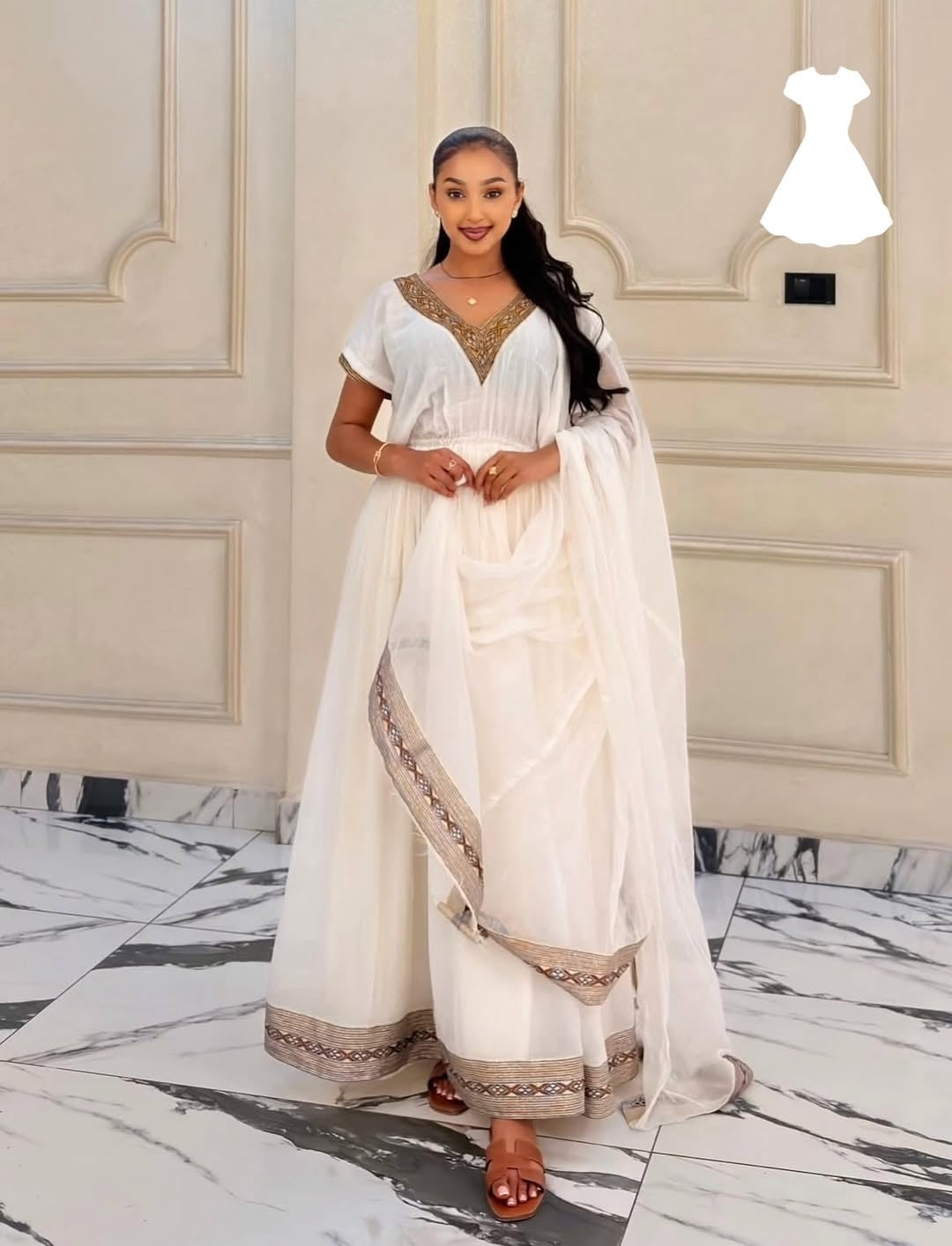 Woman in a white habesha kemis with gold details standing in a room with marble flooring and cream walls.