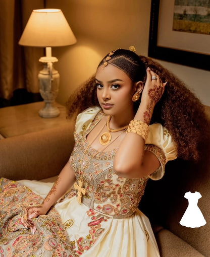 Woman in Ethiopian Wedding Dress embroidered outfit with gold jewelry in a warm-toned room.