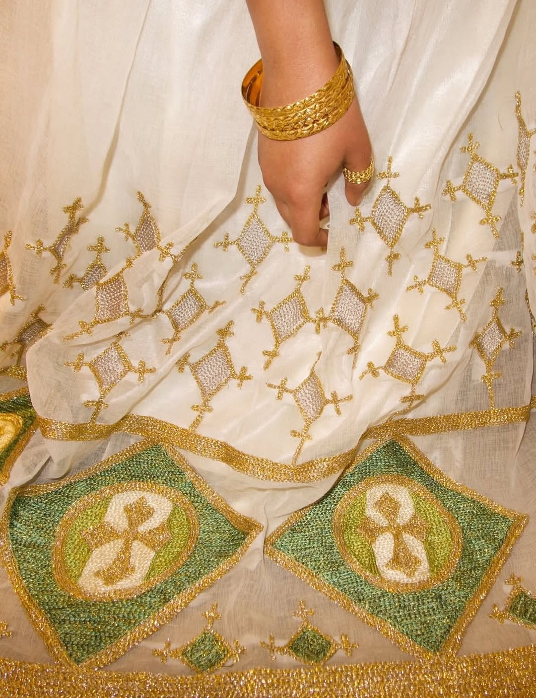 Close-up of a white fabric with gold and green Red Ethiopian Wedding Matching Set, featuring a hand wearing a gold bracelet.