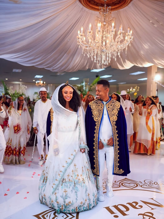 Couple in formal attire standing under a chandelier in a decorated hall with guests.