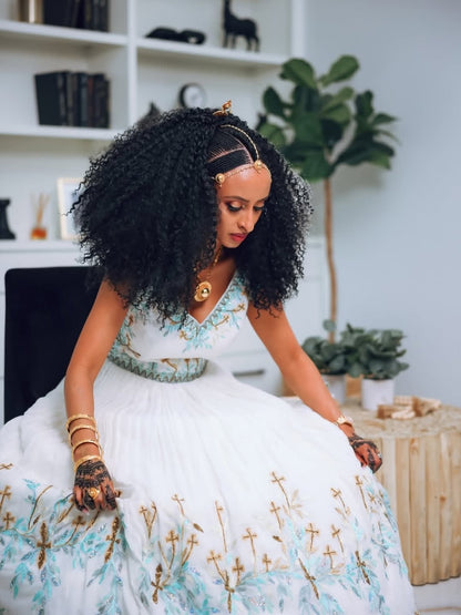 Woman in a white and blue Habesha Kemis with gold accents sitting in a room with plants and shelves.