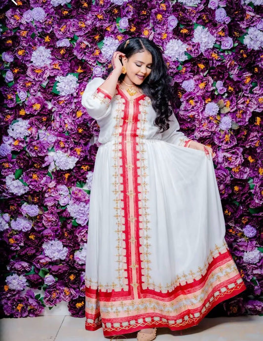 a woman wearing a traditional white Habesha kemis with vibrant red and gold embroidery running down the center and along the bottom hem. She is posing with one hand near her hair against a dense background of purple and violet flowers.
