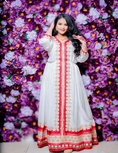 a woman wearing a traditional white Habesha kemis with vibrant red and gold embroidery running down the center and along the bottom hem. She is posing with one hand near her hair against a dense background of purple and violet flowers.
