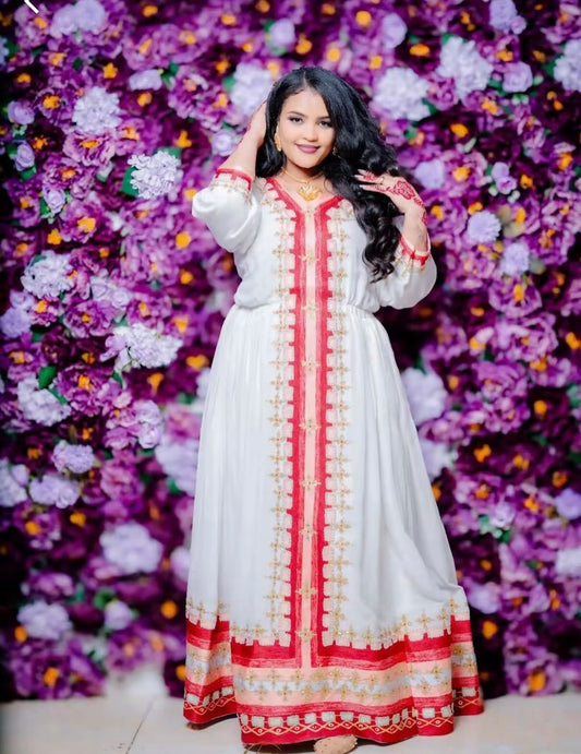 a woman wearing a traditional white Habesha kemis with vibrant red and gold embroidery running down the center and along the bottom hem. She is posing with one hand near her hair against a dense background of purple and violet flowers.
