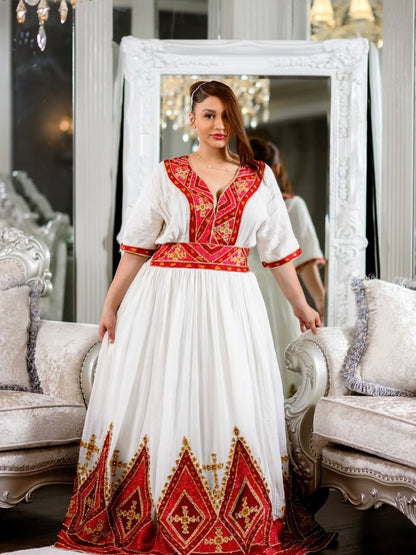 A woman wearing a traditional white and red embroidered habesha dress with gold accents, standing in a decorated room with a large ornate mirror and upholstered chairs.
