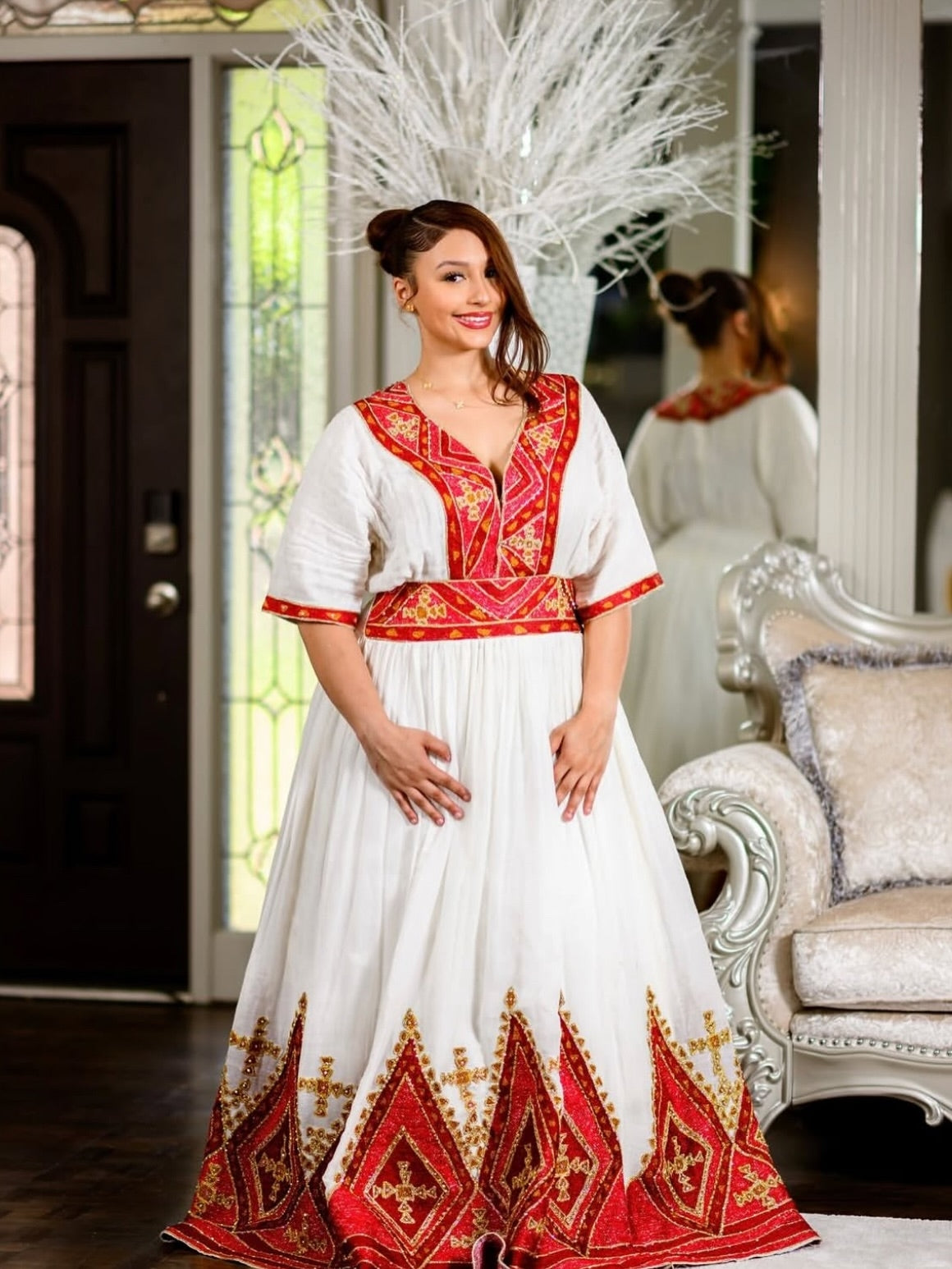 A woman wearing a traditional white habesha dress with red and gold embroidered details, standing in an elegant indoor setting with a mirror reflecting her back.