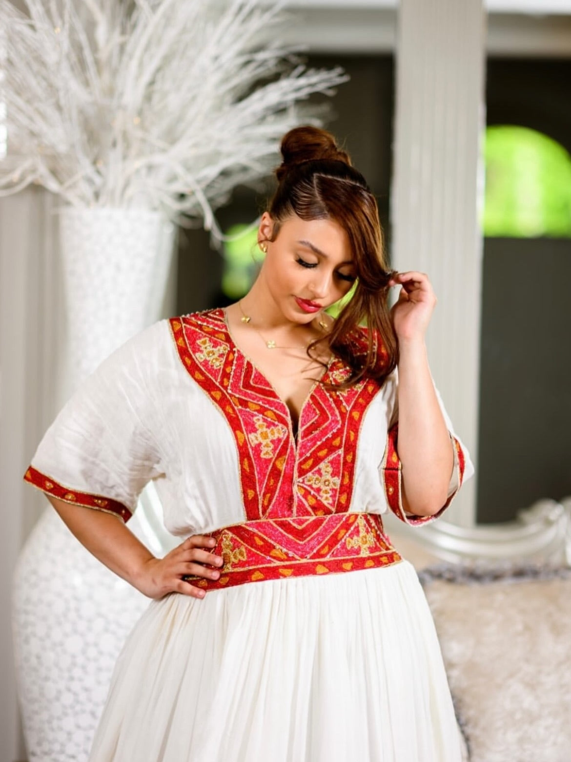 A woman wearing a traditional white and red embroidered habesha dress with gold accents, standing in a decorated room with a large ornate mirror and upholstered chairs.