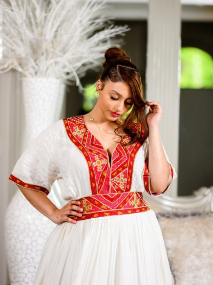 A woman wearing a traditional white and red embroidered habesha dress with gold accents, standing in a decorated room with a large ornate mirror and upholstered chairs.