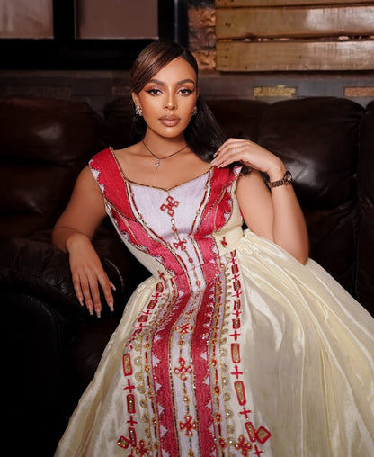 A woman modeling a traditional  Habesha Zuria in a dimly lit room with brick walls and leather sofas. The dress has a cream-colored skirt with red and gold embroidery, and a red and white embroidered bodice.