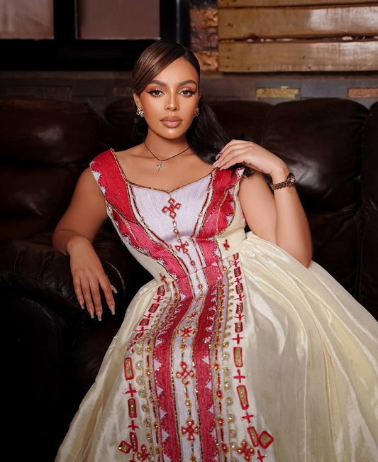 A woman modeling a traditional  Habesha Zuria in a dimly lit room with brick walls and leather sofas. The dress has a cream-colored skirt with red and gold embroidery, and a red and white embroidered bodice.
