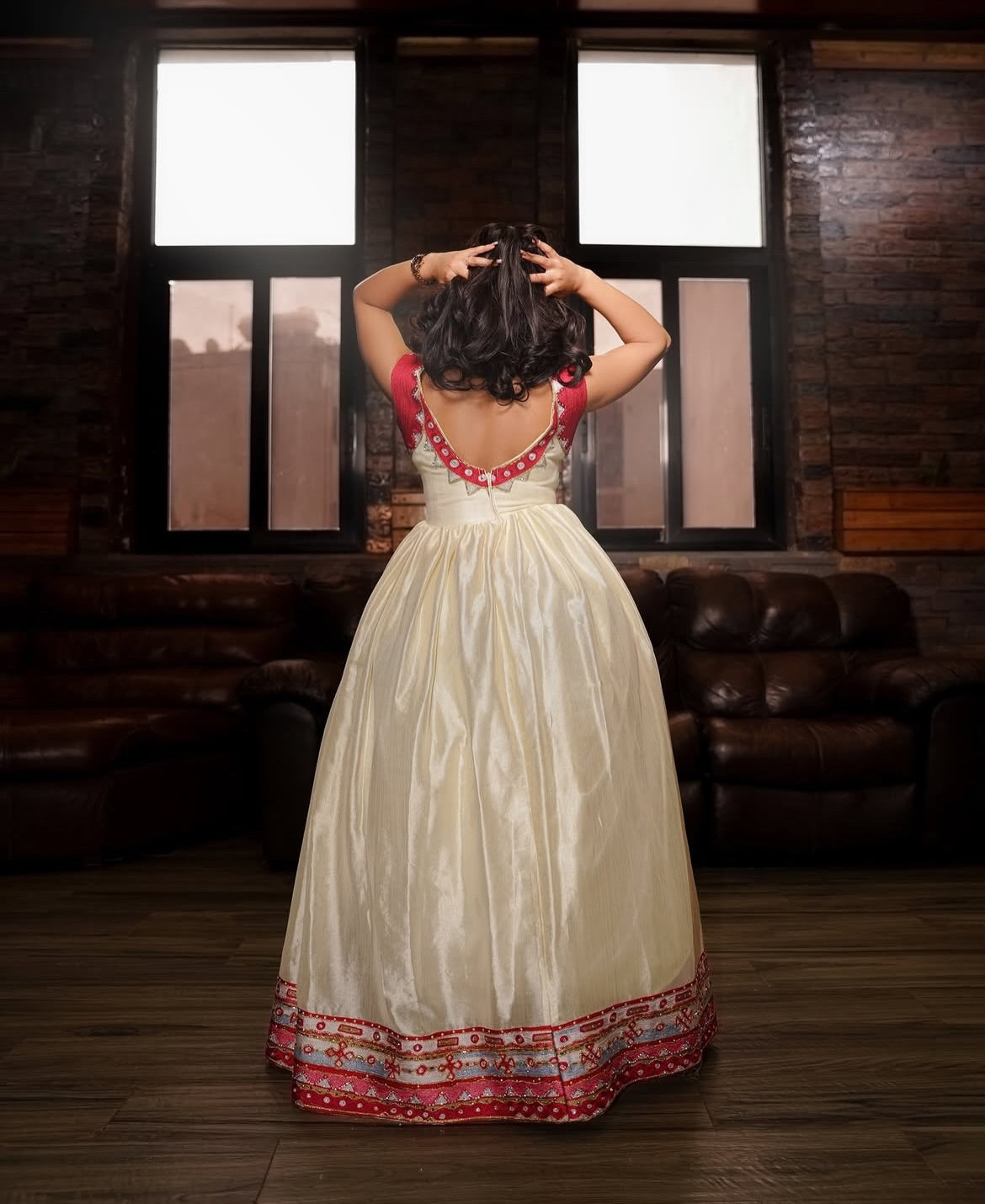 A woman stands with her back to the camera wearing a cream-colored traditional Habesha Zuria with red embroidered details on the bodice and hem, set in a dimly lit room with brick walls and leather sofas.