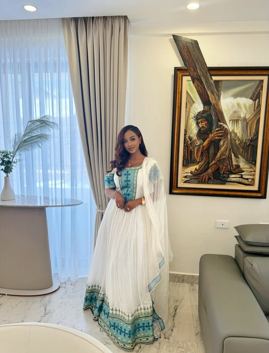 A woman wearing a traditional white and teal blue ethiopian dress with a sheer white dupatta stands in a modern living room setting with a sofa, curtains, and a framed painting on the wall.