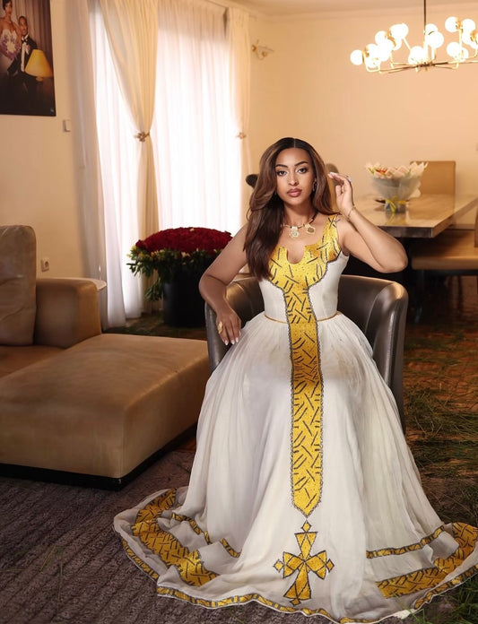 A woman wearing a white and gold embroidered Habesha Dress  sits in a chair inside a furnished room with curtains, a sofa, and a chandelier.