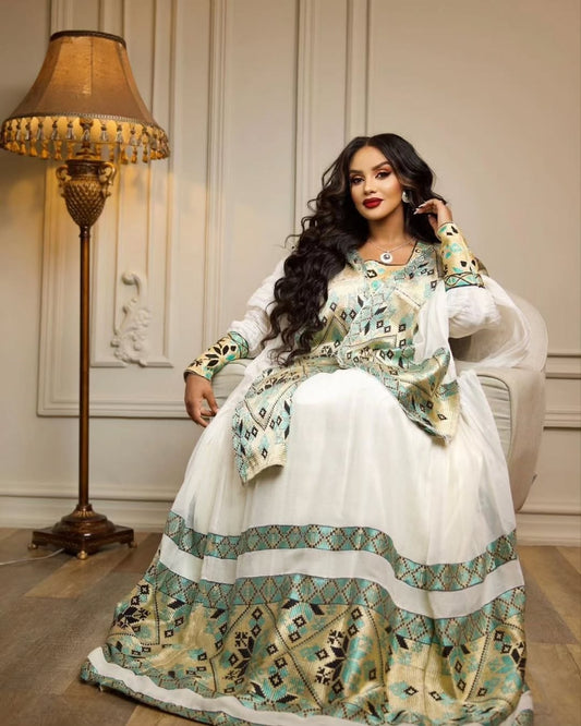 Woman in a white and green Habesha Libs  sitting in a room with a lamp and decorative wall.