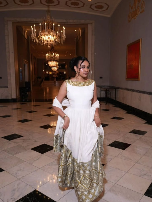 Woman in a white and gold Habesha Kemis standing in an elegant interior setting with chandeliers.