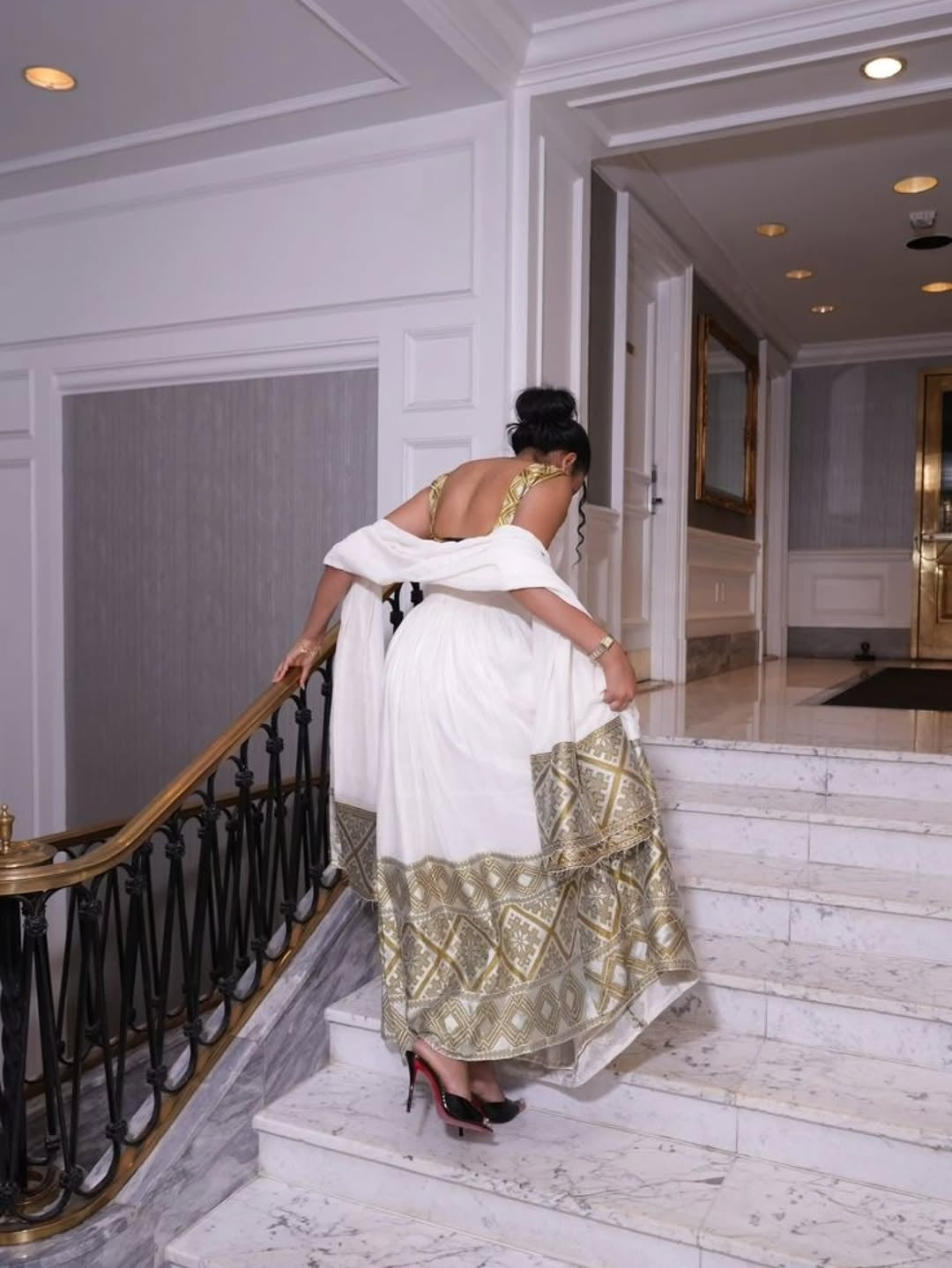 Woman in a white and gold Habesha Kemis descending a staircase in an elegant interior setting.