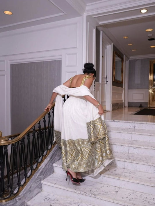 Woman in a white and gold Habesha Kemis descending a staircase in an elegant interior setting.