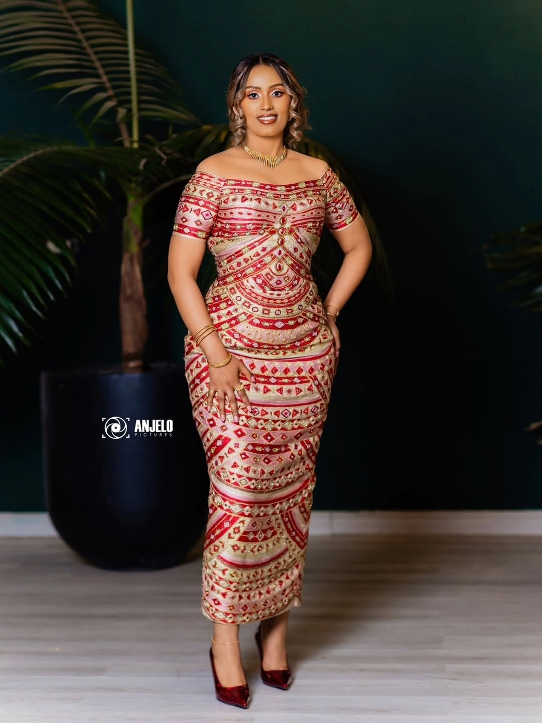 Woman in a patterned Habesha Kemis standing against a dark background with a plant.