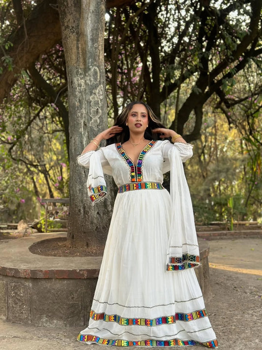 Woman in a white habesha kemis with colorful accents standing on a stone bench in a park.