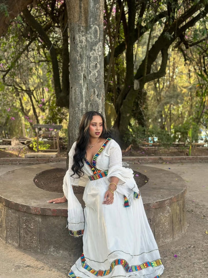 Woman in a white habesha kemis with colorful accents sitting on a stone bench in a park.