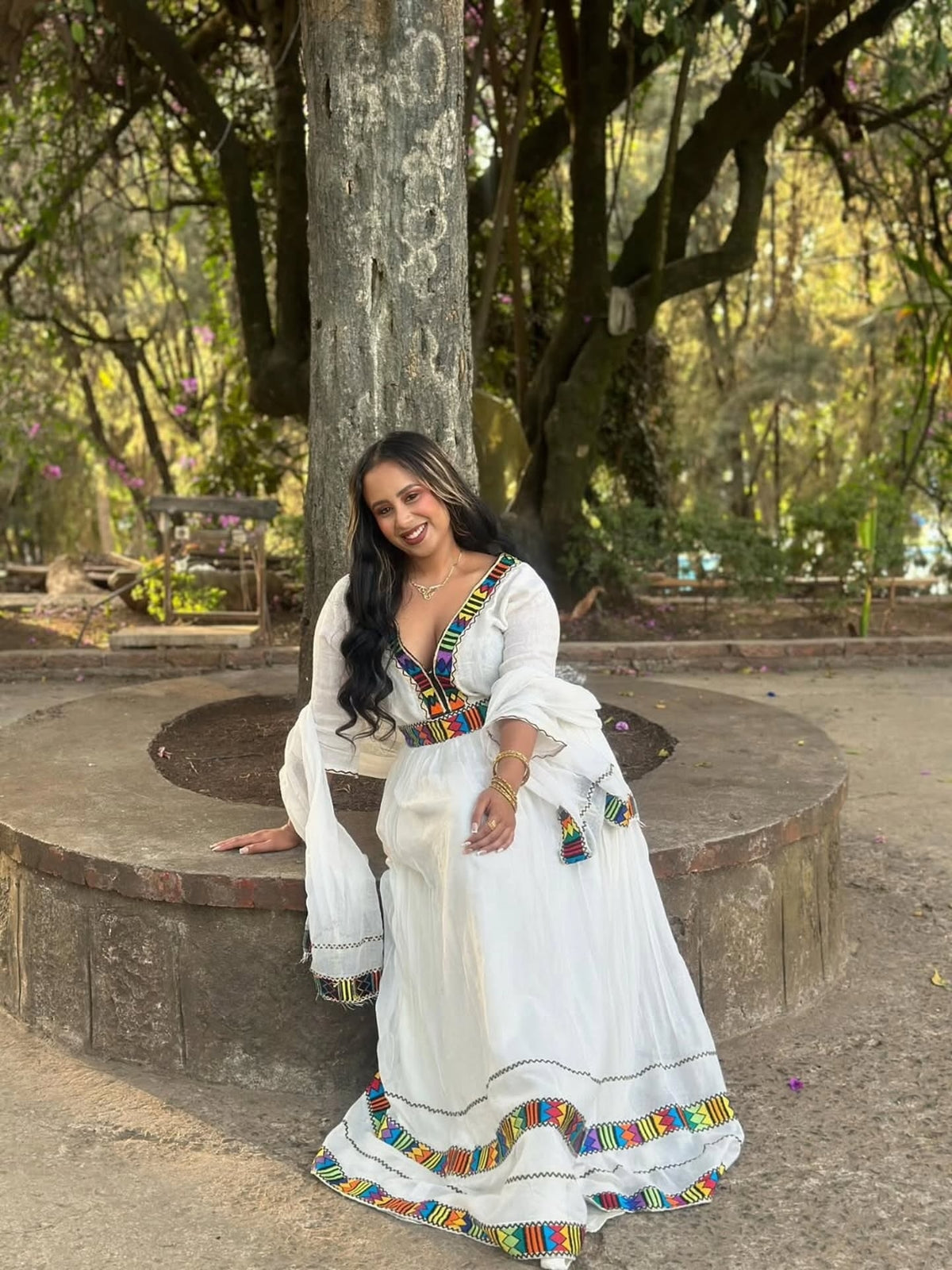 Woman in a white traditional habesha kemis with colorful patterns sitting outdoors.