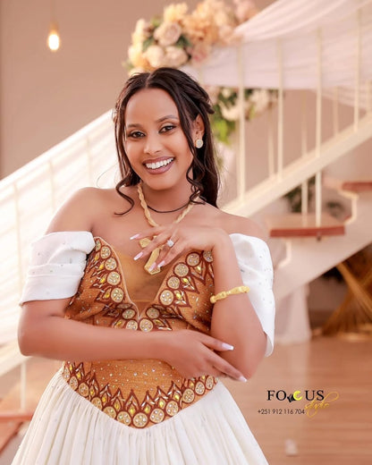 Woman in a decorated indoor setting wearing a gold and white Habesha Wedding Dress.