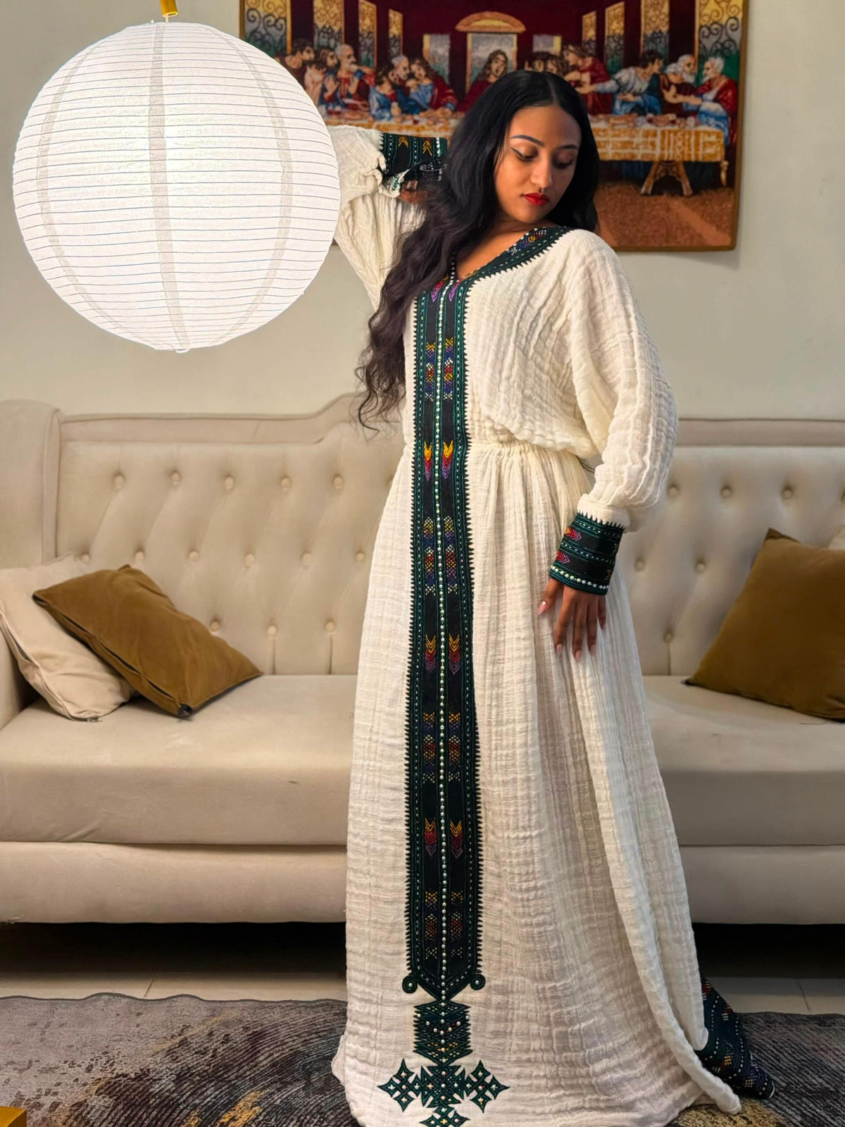 A woman wearing a long white Habesha Dress  with dark green and colorful beaded embroidery, standing in a living room in front of a cream tufted sofa and a hanging white paper lantern.
