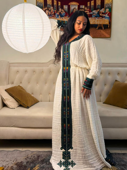 A woman wearing a long white Habesha Dress  with dark green and colorful beaded embroidery, standing in a living room in front of a cream tufted sofa and a hanging white paper lantern.