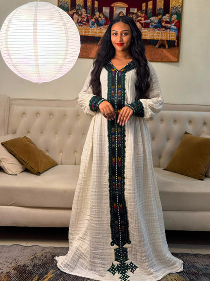 A woman wearing a long white Habesha Dress with dark green and red embroidered details, standing in a living room in front of a tufted sofa and a large hanging paper lantern.