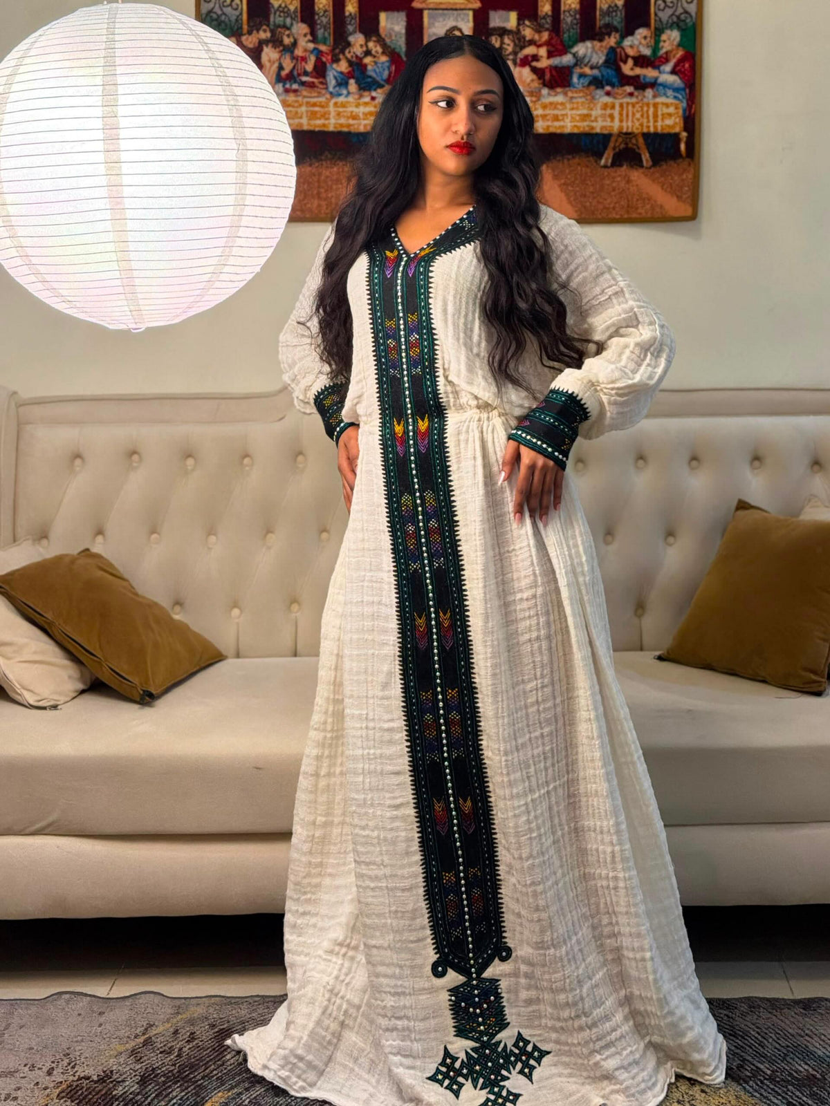 A woman wearing a long white textured kaftan Habesha Dress  with dark green and multicolor embroidered details, standing in a living room in front of a tufted sofa.