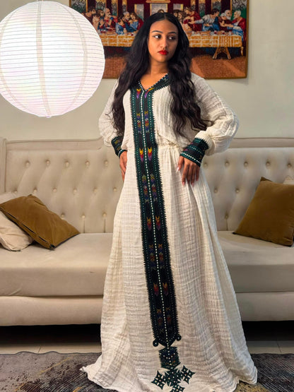 A woman wearing a long white textured kaftan Habesha Dress  with dark green and multicolor embroidered details, standing in a living room in front of a tufted sofa.