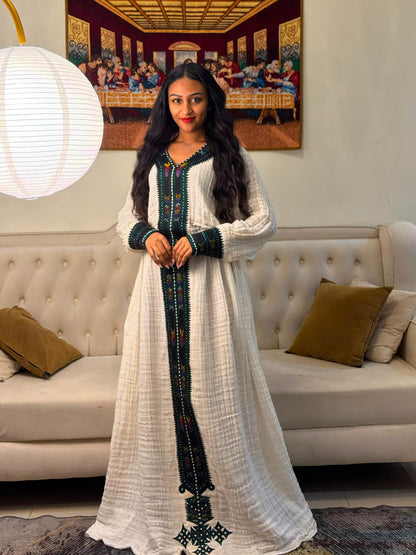 A woman stands in a living room wearing a long white textured Habesha Dress  with dark green and red embroidered details down the front and on the sleeves. A cream tufted sofa with brown pillows is behind her, along with a framed painting and a hanging white paper lantern.