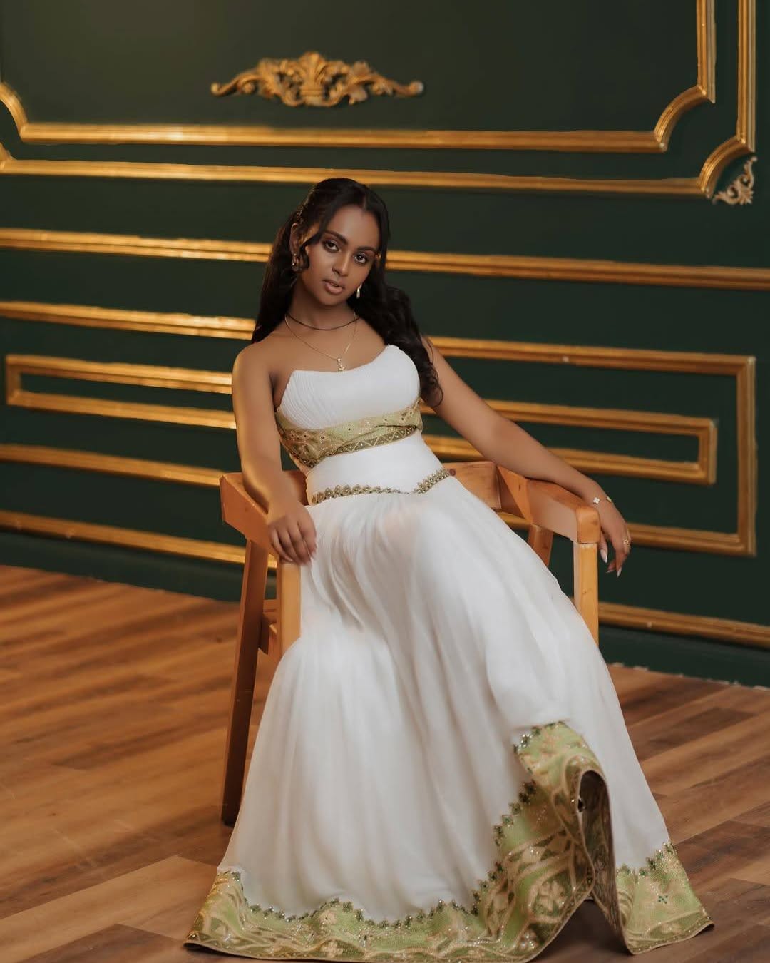 A woman wearing a white strapless Eritrean Zuria with gold detailing sits on a wooden chair in front of a dark green wall with gold trim.