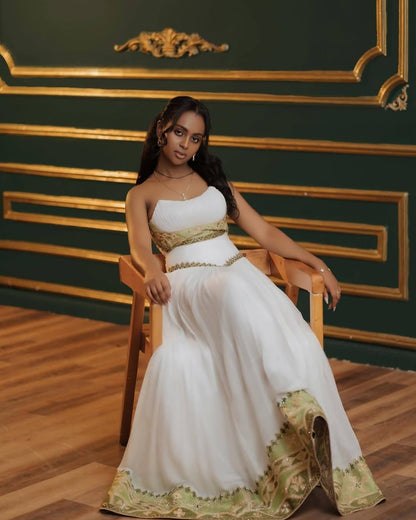 A woman wearing a white strapless Eritrean Zuria with gold detailing sits on a wooden chair in front of a dark green wall with gold trim.