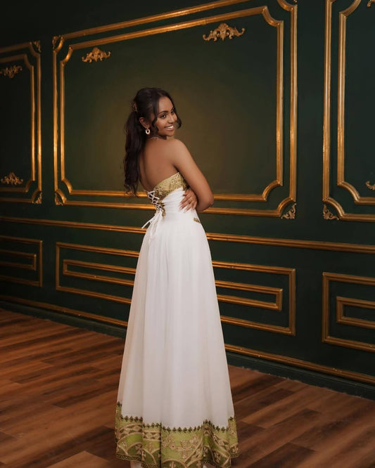 A woman wearing a strapless white formal Eritrean Zuria with gold trim and a matching gold patterned hem, standing in front of an ornate dark green wall with gold paneling.
