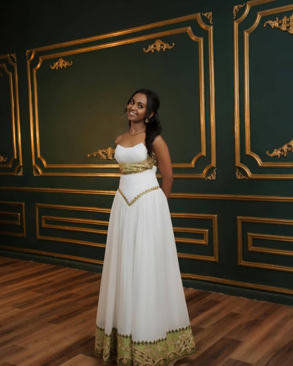 A woman wearing a white strapless Eritrean Zuria with gold embroidery and a gold waist sash, standing in front of a dark green wall with gold paneling.
