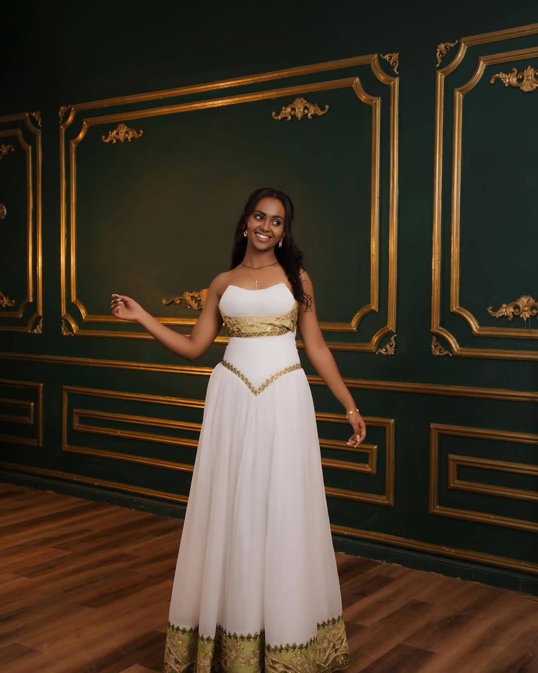 A woman modeling a white strapless maxi Eritrean Zuria with gold trim, standing in front of a dark green wall with gold paneling and a wooden floor.