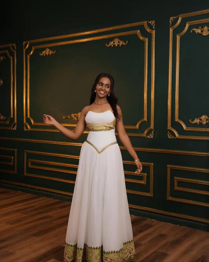 A woman modeling a white strapless maxi Eritrean Zuria with gold trim, standing in front of a dark green wall with gold paneling and a wooden floor.