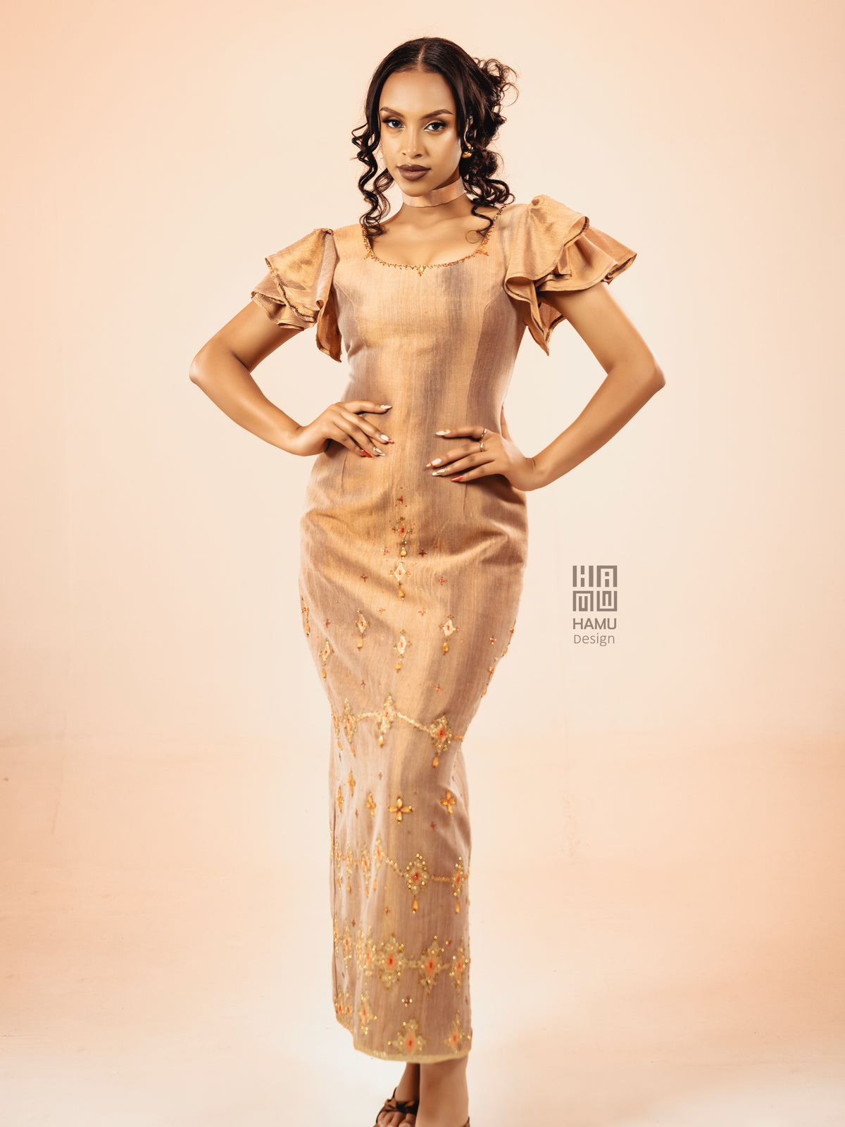 A woman modeling a beige maxi Habesha Kemis with ruffled short sleeves and gold floral embroidery, posed against a solid light peach studio background.
