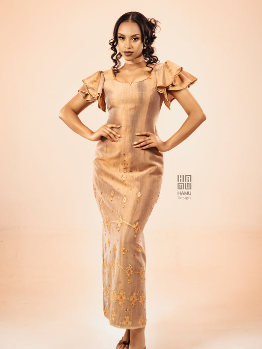 A woman modeling a beige maxi Habesha Kemis with ruffled short sleeves and gold floral embroidery, posed against a solid light peach studio background.