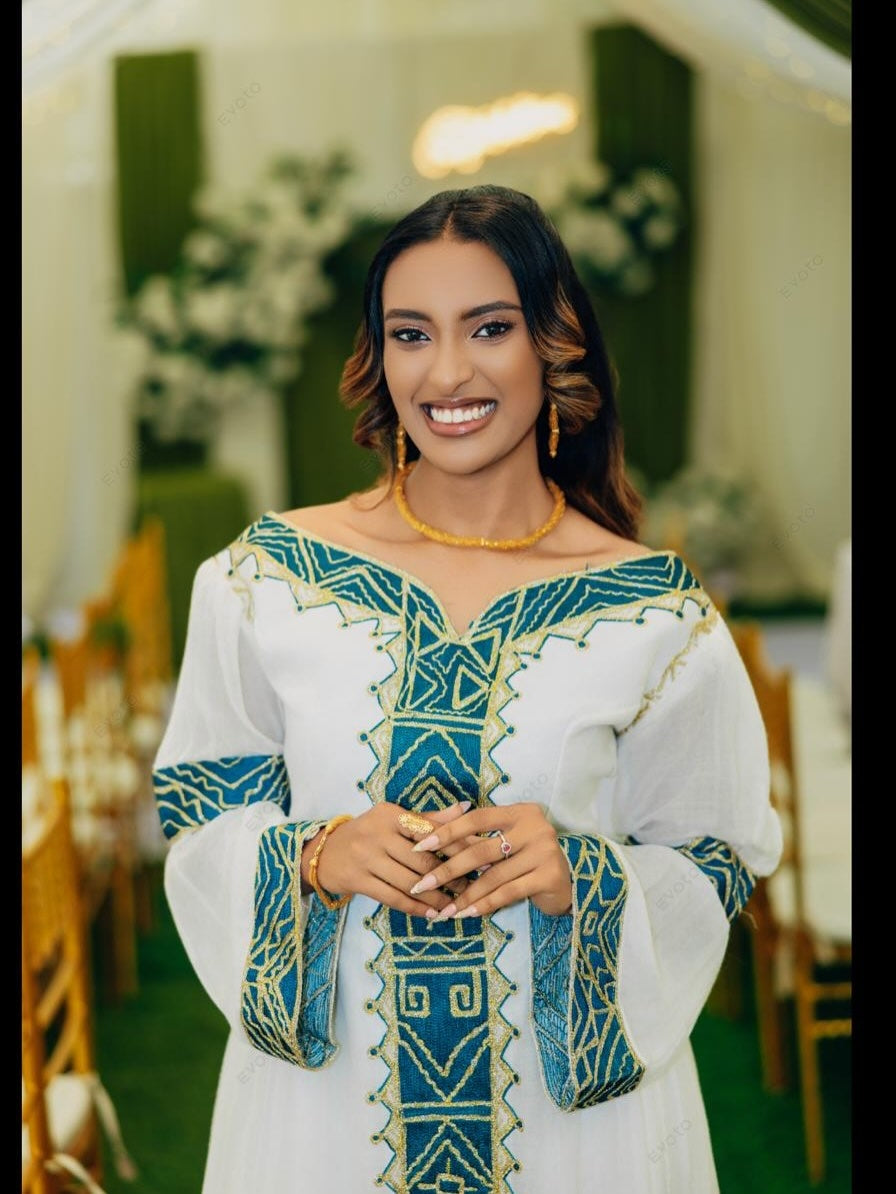 A smiling woman wearing a white off-the-shoulder traditional Habesha zuria with teal and gold geometric embroidery, standing in a decorated event space with blurred chairs and greenery in the background.
