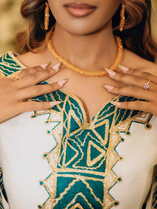 A close-up lifestyle shot of a woman wearing a white Habesha zuria with a deep V-neckline, heavily decorated with teal and gold geometric embroidery. She is also wearing a gold beaded necklace and matching gold hoop earrings.