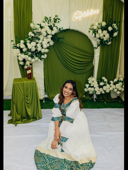 A woman wearing a traditional white Habesha zuria with green and teal embroidery sits in front of a decorated green and white event backdrop with floral arrangements and a lit 'Congratulations' sign.