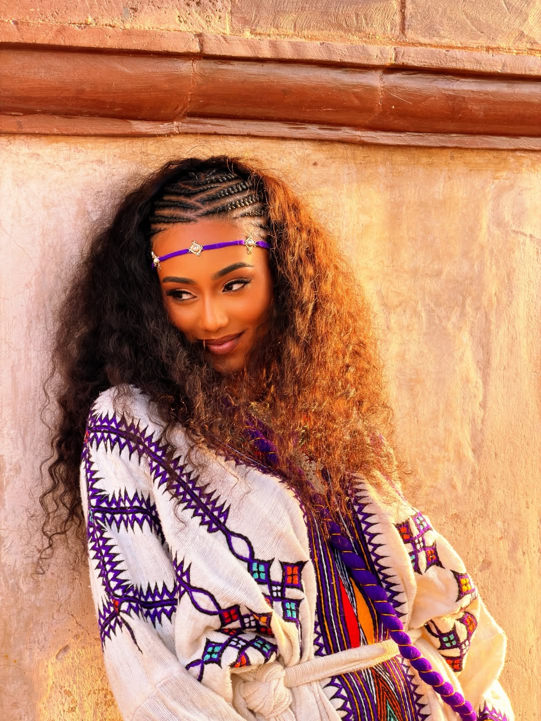 Woman with braided hair wearing a colorful patterned gonder fetel against a textured wall.