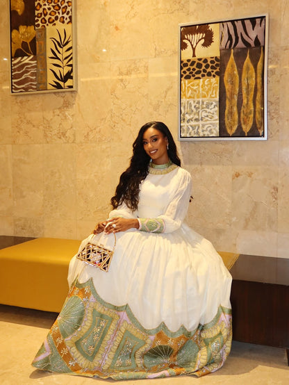 Woman in a white Habesha Dress with colorful patterns sitting on a bench in a room with decorative wall art.