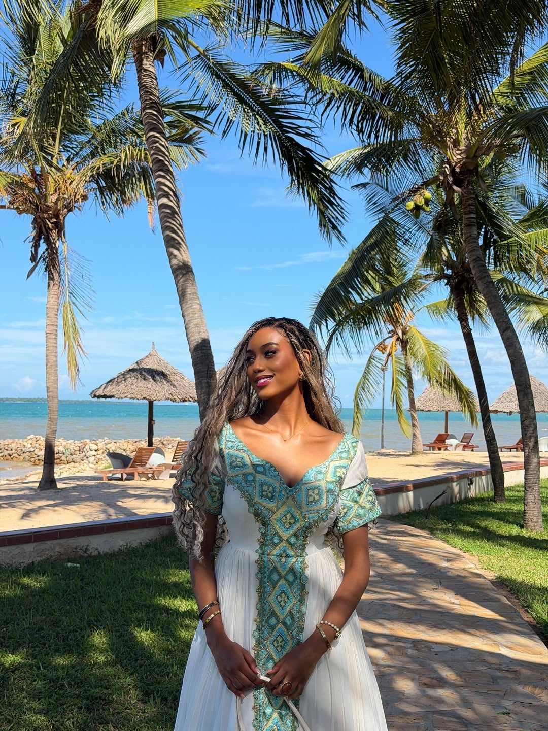 Woman in a white and green Green Eritrean Libs standing on a tropical beach with palm trees and clear blue water.