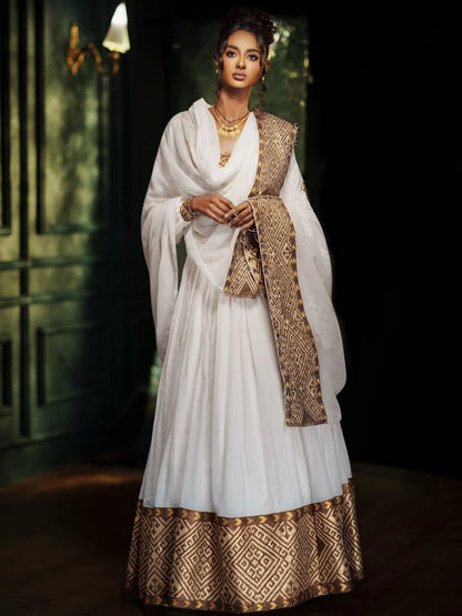 A woman models a traditional white Ethiopian Traditional Clothing with gold patterned borders and a matching draped shawl in a dimly lit interior setting.