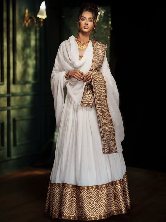 A woman models a traditional white Ethiopian Traditional Clothing with gold patterned borders and a matching draped shawl in a dimly lit interior setting.