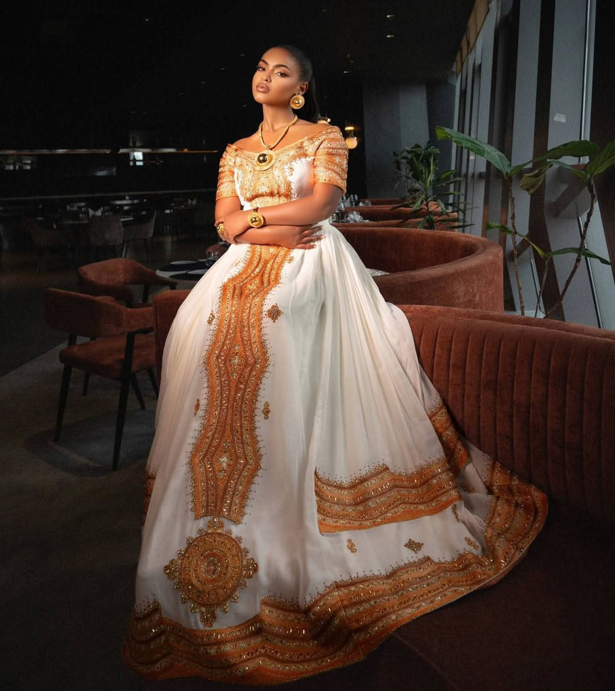 A woman wearing a traditional South Asian ball Ethiopian Store  with white fabric and intricate gold embroidery, posing in a dimly lit indoor setting with velvet seating.